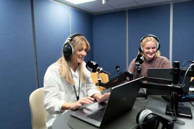 To kvinner sitter ved et bord i et studio for lydopptak. De har på hodetelefoner, og foran seg har de hver sin mikrofon og pc.