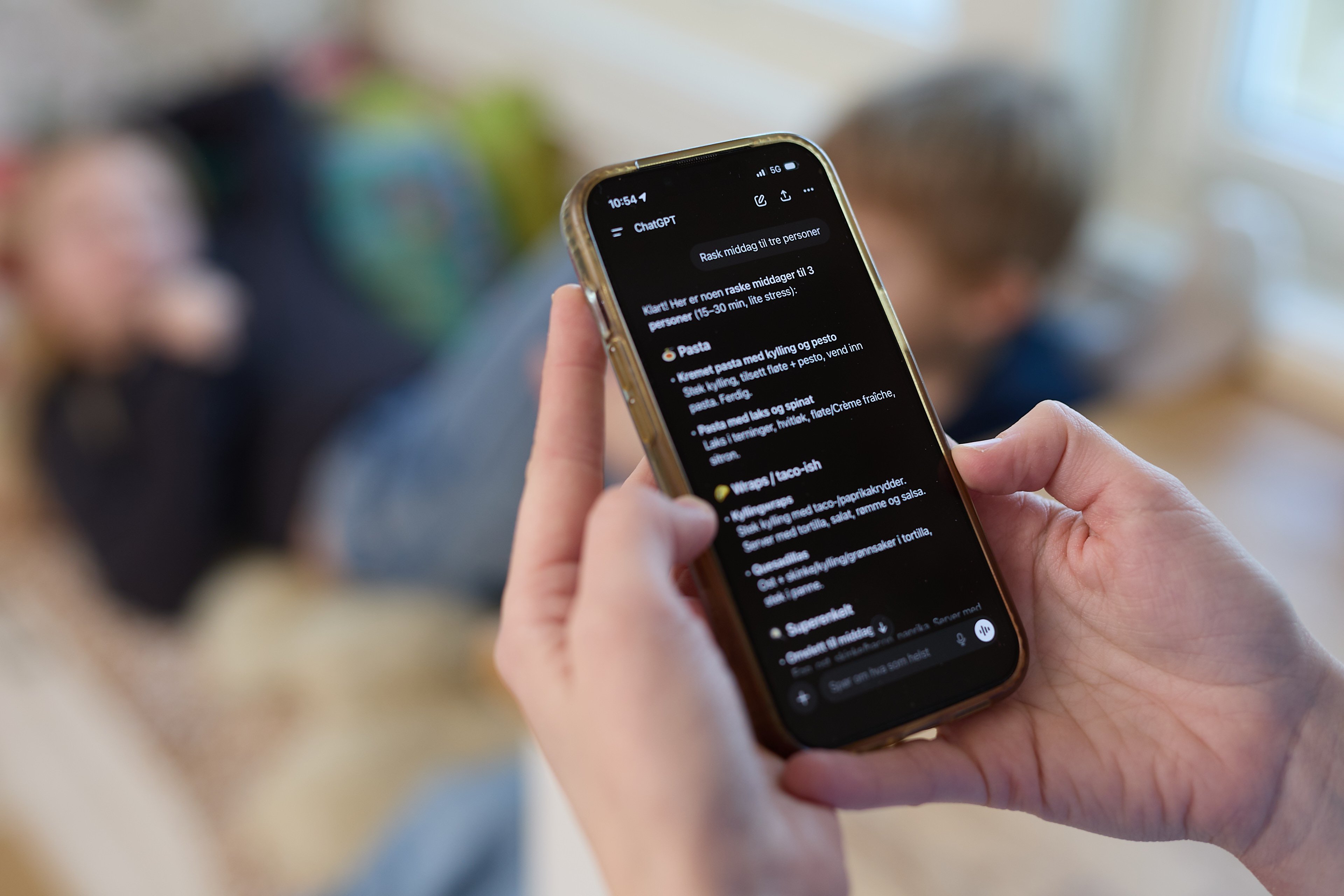 En person holder en mobiltelefon og leser en samtale med ChatGPT på skjermen. I bakgrunnen, ute av fokus, sitter to personer i en sofa i en stue.