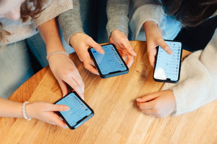 Tre personer sitter rundt et bord og holder hver sin mobiltelefon. Skjermene viser meldingsapper, og hendene deres er i fokus.