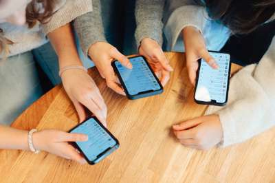 Tre personer sitter rundt et bord og holder hver sin mobiltelefon. Skjermene viser meldingsapper, og hendene deres er i fokus.