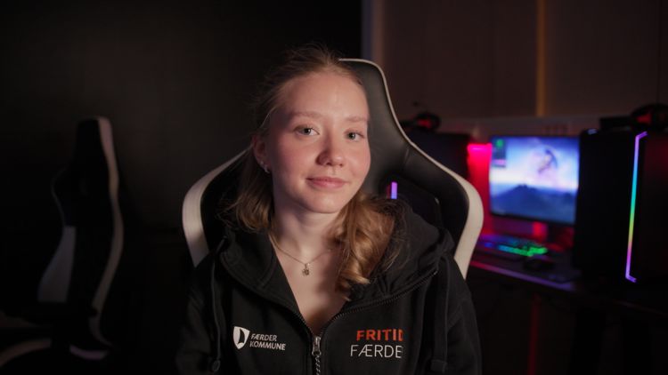 Ung kvinne sitter i en gamingstol i et mørkt rom med datamaskiner i bakgrunnen. Hun har på seg en hettegenser med teksten «Fritid Færder» og «Færder kommune».