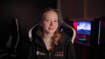 Ung kvinne sitter i en gamingstol i et mørkt rom med datamaskiner i bakgrunnen. Hun har på seg en hettegenser med teksten «Fritid Færder» og «Færder kommune».