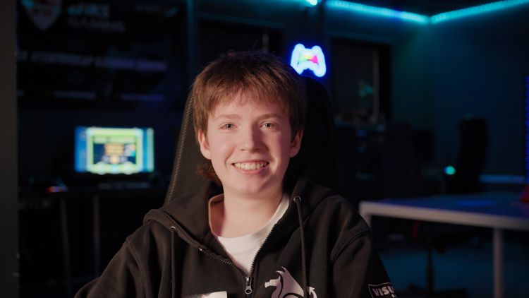 Ung gutt sitter i en gamingstol i et rom med blå belysning og datamaskiner i bakgrunnen. Han smiler og har på seg en hettegenser med logoer.