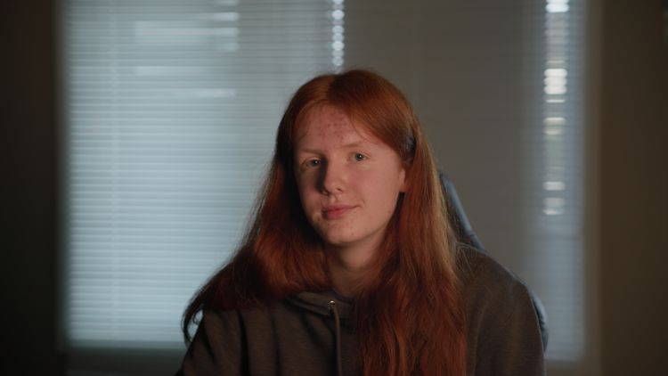 En ung guttmed langt rødt hår sitter i en gamingstol foran et vindu med persienner. Personen har på seg en mørk hettegenser og ser rolig mot kamera.