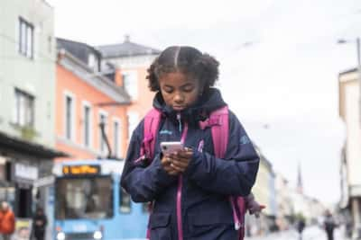 Jente på cirka ti år med skolesekk på ryggen går i en bygate og ser på telefonen sin.