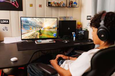 En gutt tidlig i tenårene sitter tilbakelent i gamingstol og spiller. Han er avbildet på skrå bakfra. Han har hvit t-skjorte og mørkt hår. I hendene har han en kontroller og skjermen er på en pult foran ham.