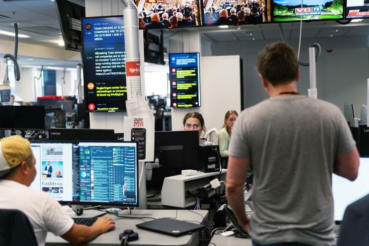 Arbeid ved desken i en avisredaksjon. Tre personer sitter ved hver sin pult mens en fjerde står med ryggen til fotografen ved pulten nærmest.