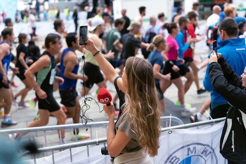 Ei kvinneleg journalist holder høyre arm i veret for å filme med telefonen sin. I venstre hand har ho ein mikrofon, og det heng eit kamera rundt halsen. I bakgrunnen løper mange mennesker forbi med startnummer på brystet.
