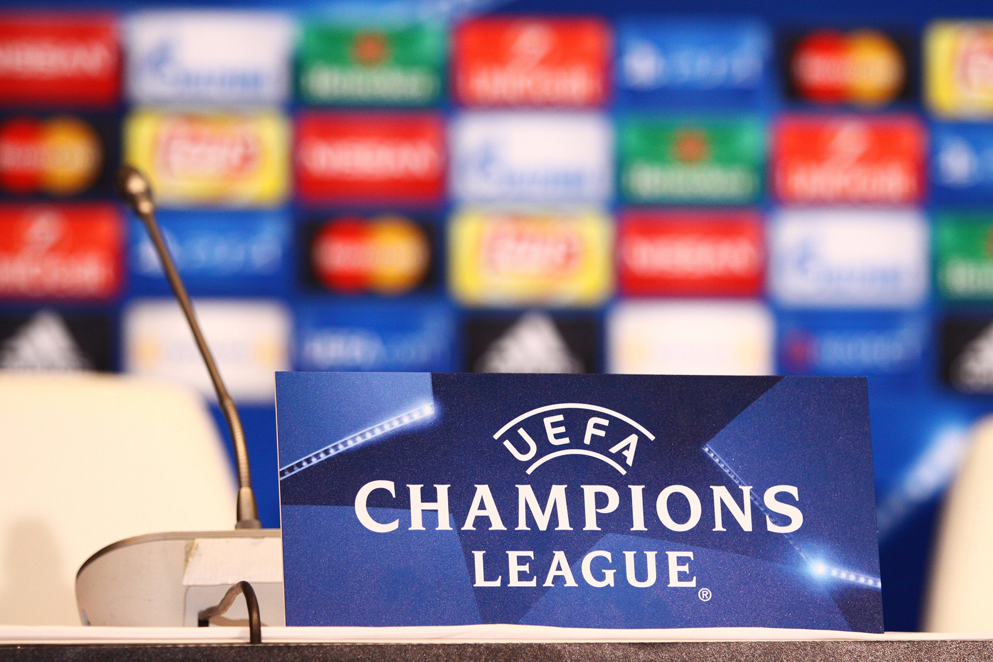 Podium med mikrofon foran et skilt med teksten «UEFA Champions League». I bakgrunnen sees en uskarp vegg med fargerike sponsorlogoer, typisk fra en pressekonferanse i fotball.