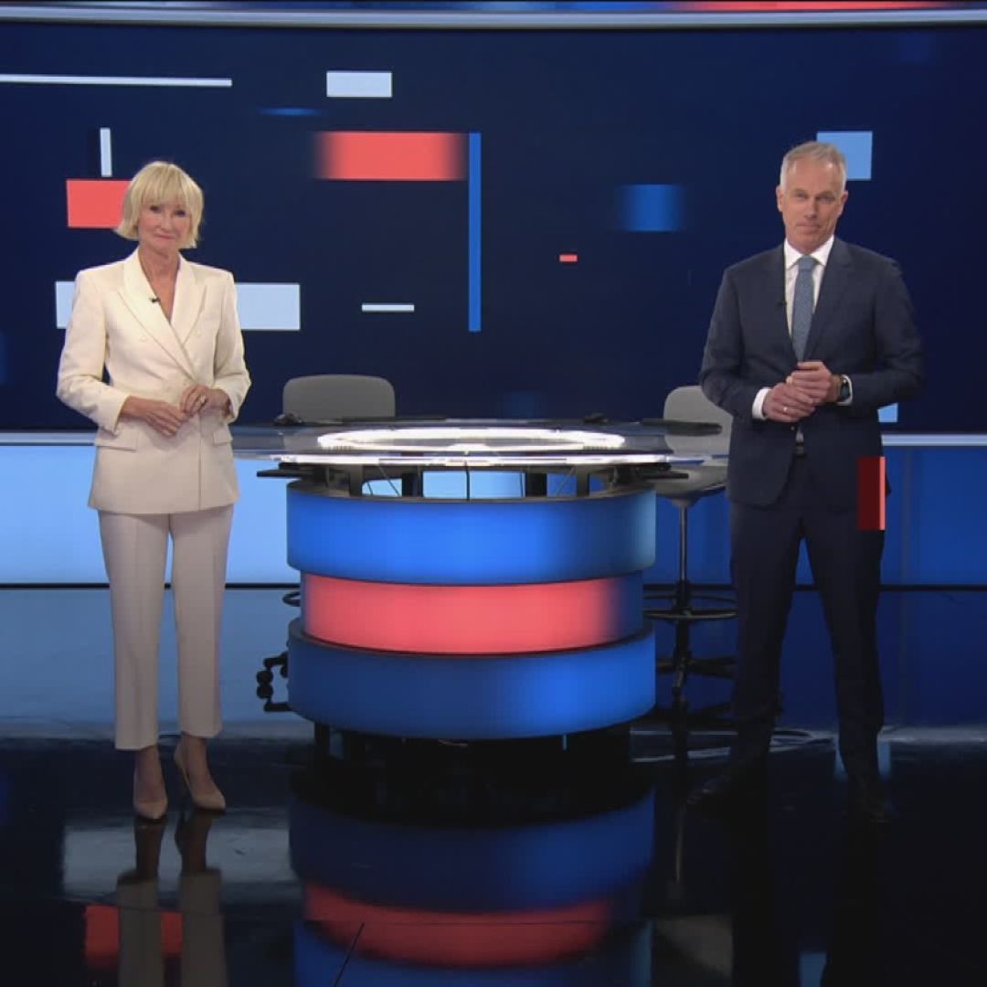 To nyhetsankere st&aring;r p&aring; hver sin side av et rundt nyhetsbord med bl&aring;tt og r&oslash;dt lys, foran en stor skjerm med abstrakte bl&aring;, r&oslash;de og hvite grafiske elementer. Foto.