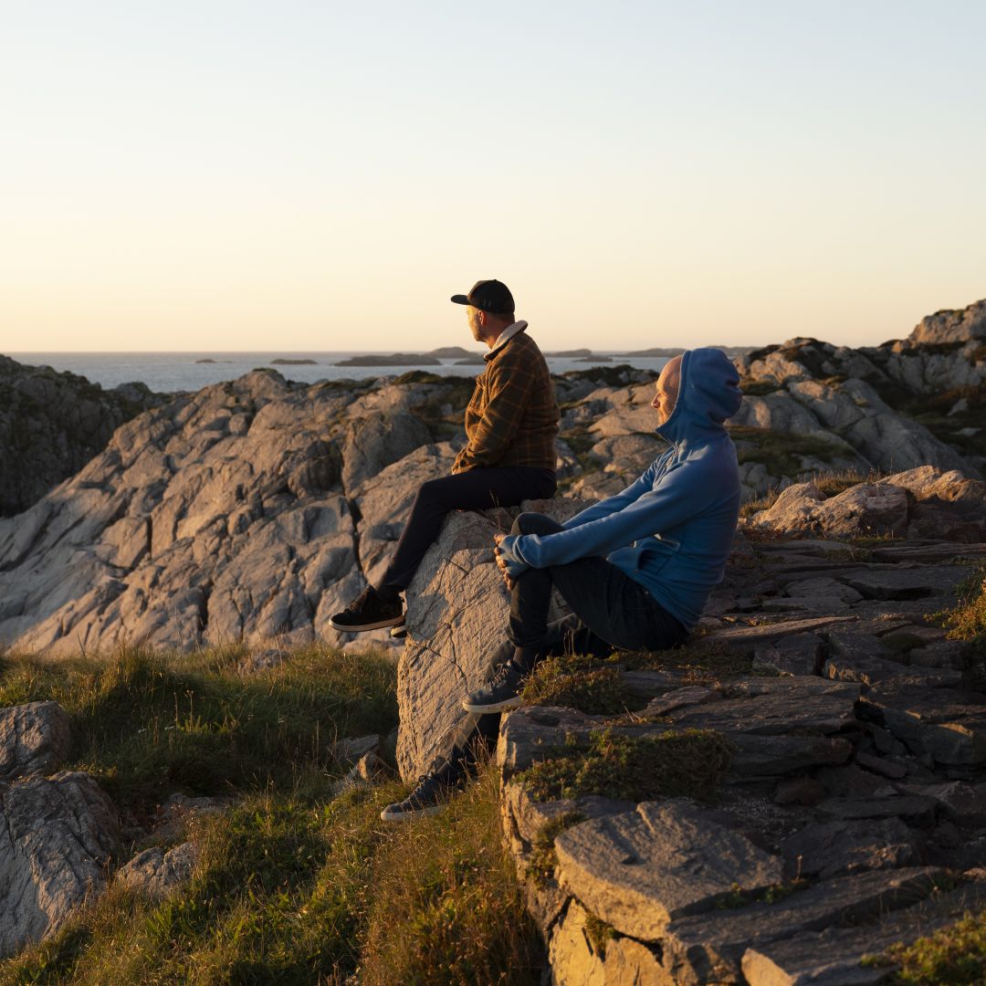 To personer sitter p&aring; svaberg ved kysten i lav kveldssol og ser utover havet og sm&aring; &oslash;yer i horisonten.